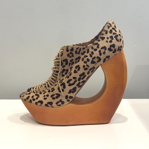 May include: A unique platform shoe with a leopard print upper and a wooden, open-circle heel. The shoe features a lace-up design and a light brown wooden platform. The heel is approximately 6 inches high.