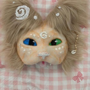 Può includere: Una maschera da gatto fatta a mano con un viso color beige, occhi blu e verdi e soffice pelliccia beige. La maschera ha dettagli bianchi e una piccola lingua rosa. Una catena d'oro con un ciondolo a conchiglia pende da un lato. La maschera è su uno sfondo a scacchi rosa e bianco.