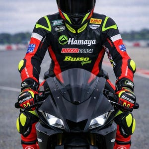 May include: A person in a black, red, and yellow motorcycle racing suit and helmet sits on a black sport motorcycle. The suit has "Hamaya" and "Rosso Corsa" logos. The helmet is black with red and yellow accents.