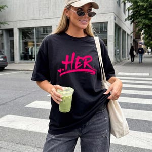 Puede incluir: Camiseta negra con la palabra "HER" en rosa brillante, con dos pequeños corazones debajo. La camiseta es de estilo casual. La persona lleva una gorra beige y gafas de sol.