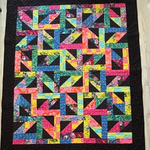 May include: A rectangular quilt featuring a geometric pattern of multi-colored fabric strips in shades of pink, yellow, blue, and green, arranged in a repeating block design. The quilt is bordered by a solid black fabric with a swirling pattern.