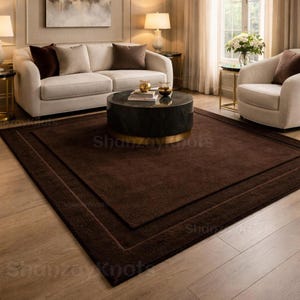 May include: A dark brown rectangular rug with a bordered design, placed on a hardwood floor. The rug is in a living room setting, with a white sofa, chairs, and a coffee table. The room is well-lit with lamps and natural light.