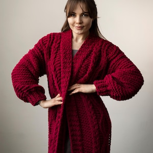 Puede incluir: Cárdigan largo de punto color burdeos con diseño de punto trenzado. El cárdigan tiene cuello chal y mangas largas. La prenda está hecha de un hilo grueso y voluminoso y llega hasta la rodilla.