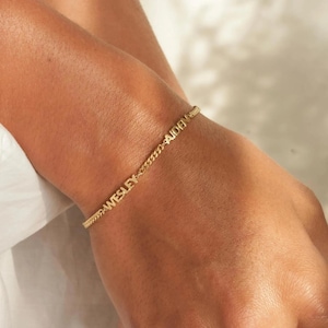 May include: A gold-colored chain bracelet with the names "WESLEY" and "AIDEN" spelled out in capital letters. The bracelet is worn on a wrist and has a delicate, textured chain design.