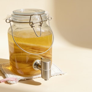 Könnte beinhalten: Ein Getränkespender aus klarem Glas mit einem Metalldrahtbügelverschluss und einem Zapfhahn aus Edelstahl. Das Glas ist mit einer goldgelben Flüssigkeit gefüllt, in der sich helle organische Stoffe befinden, und steht auf einem weißen Stoff.