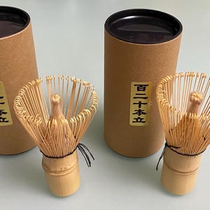 以下が含まれることがあります： 細く湾曲した穂先と中央の持ち手を持つ2本の竹製抹茶用茶筅。それぞれに茶色の円筒形紙製収納ケースが付属している。各ケースには、黒い日本語の文字「百二十本立」が印刷された金色の長方形のラベルが貼られている。