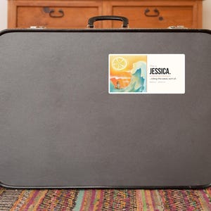 May include: A rectangular sticker featuring a graphic of a sun, a large ocean wave, and a person, with the text "This is JESSICA. ...riding the wave. sort of." The sticker is applied to the centre of a dark grey, hard-shell suitcase with a black handle.