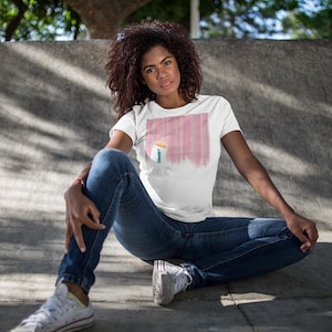 May include: White t-shirt with a pink and white vertical stripe design. The shirt is paired with blue jeans and white sneakers. The person is sitting on a concrete surface.