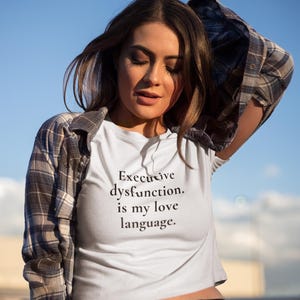 Puede incluir: Top corto blanco con el texto "Executive dysfunction. is my love language." La camiseta se combina con pantalones negros y una camisa a cuadros. El fondo es un cielo azul claro.