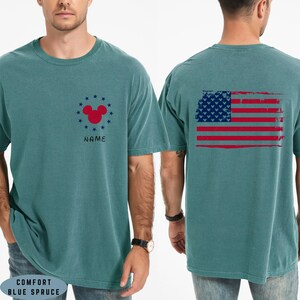 Op de afbeelding: Comfort Blue Spruce T-shirt met een verweerd Amerikaans vlagontwerp op de rug. De voorkant heeft een rode Mickey Mouse-kop omringd door blauwe sterren en het woord "NAME".