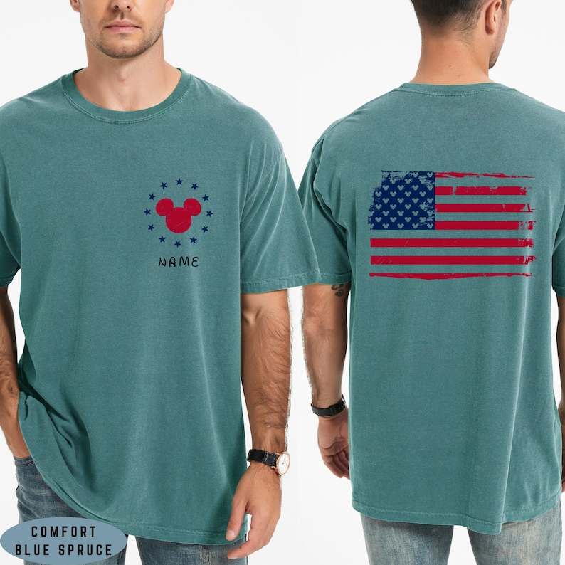 Op de afbeelding: Comfort Blue Spruce T-shirt met een verweerd Amerikaans vlagontwerp op de rug. De voorkant heeft een rode Mickey Mouse-kop omringd door blauwe sterren en het woord "NAME".