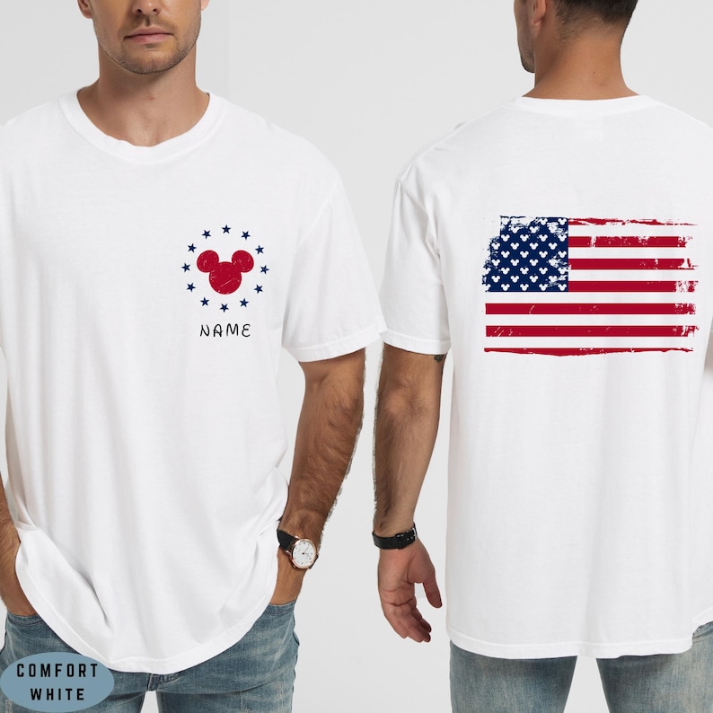 Op de afbeelding: Wit T-shirt met een rood Mickey Mouse hoofd ontwerp omringd door blauwe sterren, met het woord "NAME" eronder. De achterkant van het shirt toont een verweerde Amerikaanse vlag. Het shirt is comfort wit van kleur.