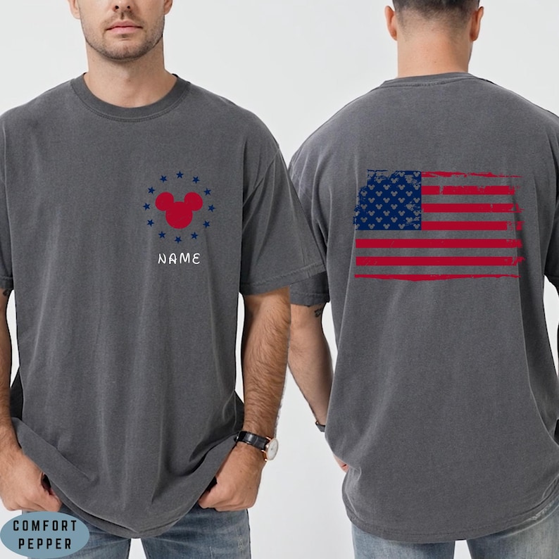 Op de afbeelding: Grijs T-shirt met een rode Mickey Mouse-kop omringd door blauwe sterren en het woord "NAME". De achterkant van het shirt toont een verweerd Amerikaans vlagontwerp. Het shirt is gemaakt van een zacht, comfortabel materiaal.