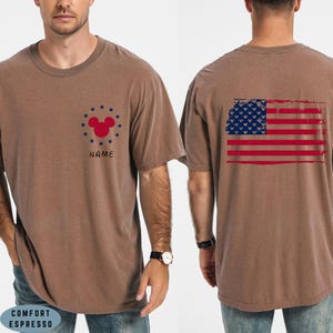 Op de afbeelding: Bruin t-shirt met een verweerde Amerikaanse vlag op de rug. De voorkant heeft een rode Mickey Mouse-kop omringd door blauwe sterren en het woord "NAME". Het shirt is gelabeld "COMFORT ESPRESSO".