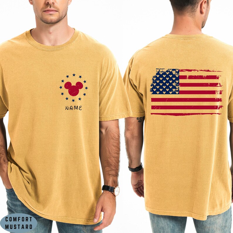 Op de afbeelding: Mosterdkleurig T-shirt met een rood Mickey Mouse-hoofdontwerp omringd door sterren en het woord "NAME". De achterkant van het shirt toont een versleten Amerikaans vlagontwerp met Mickey Mouse-hoofden in het blauwe veld.