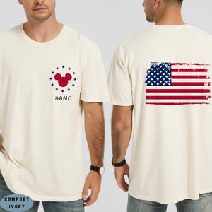 Op de afbeelding: Cr&egrave;mekleurig T-shirt met een rood Mickey Mouse hoofd ontwerp omringd door blauwe sterren en het woord "NAME" in zwart. De achterkant van het shirt toont een distressed Amerikaanse vlag in rood, wit en blauw.