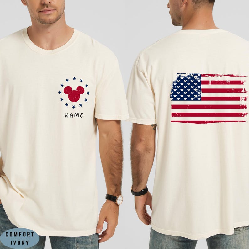 Op de afbeelding: Cr&egrave;mekleurig T-shirt met een rood Mickey Mouse hoofd ontwerp omringd door blauwe sterren en het woord "NAME" in zwart. De achterkant van het shirt toont een distressed Amerikaanse vlag in rood, wit en blauw.