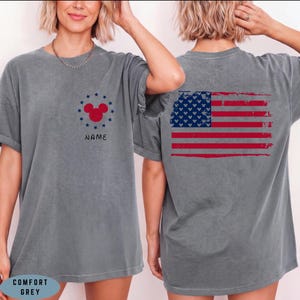 Op de afbeelding: Comfort grijs t-shirt met een verweerd Amerikaans vlagontwerp op de rug. De voorkant heeft een rode Mickey Mouse-kop omringd door blauwe sterren en het woord "NAME". Het shirt is gemaakt van een zacht materiaal.