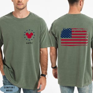 Op de afbeelding: Olijfgroen t-shirt met een verweerd Amerikaans vlagontwerp op de rug. De voorkant heeft een rode Mickey Mouse-kop omringd door blauwe sterren en het woord "NAME". Gemaakt van Comfort Moss materiaal.