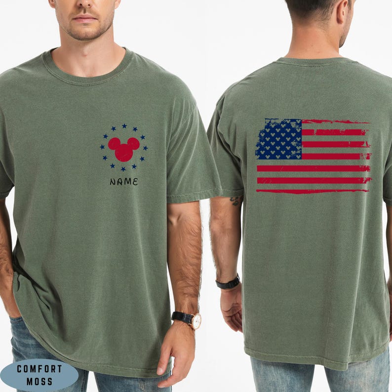 Op de afbeelding: Olijfgroen t-shirt met een verweerd Amerikaans vlagontwerp op de rug. De voorkant heeft een rode Mickey Mouse-kop omringd door blauwe sterren en het woord "NAME". Gemaakt van Comfort Moss materiaal.