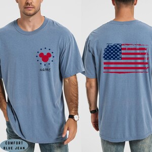 Op de afbeelding: Comfort blauw spijkerbroek t-shirt met een verweerde Amerikaanse vlag op de rug. De voorkant heeft een rood Mickey Mouse hoofd ontwerp omringd door blauwe sterren en het woord "NAME".