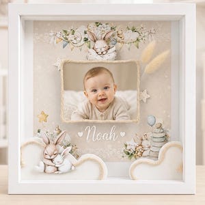 Può includere: Scatola ricordo per bambini con cornice bianca con foto di un bambino sorridente. La scatola presenta un tema coniglietto con elementi decorativi, tra cui figure di coniglietti, nuvole e il nome "Noah". Lo sfondo è beige chiaro con stelle.