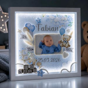 Op de afbeelding: Een vierkante witte schaduwdooslijst met een verlichte baby-herinneringsdisplay in blauwtinten. Het bevat een centrale babyfoto, de naam Fabian, de datum 25.03.2026, geboortegegevens van 55cm en 4002g, teddybeerillustraties en houten blokken met de tekst OH BABY.