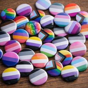 Può includere: Una collezione di bottoni rotondi colorati con vari disegni di bandiere del Pride LGBTQ+. Alcuni bottoni presentano strisce arcobaleno, mentre altri mostrano il testo "Love is Love". I bottoni sono disposti su una superficie di legno.