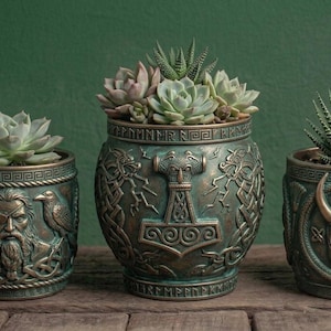 May include: Three cylindrical planters with a metallic bronze and green finish. The left planter features a bearded figure with two ravens. The center planter displays a hammer symbol. The right planter shows a horned helmet encircled by a serpent.