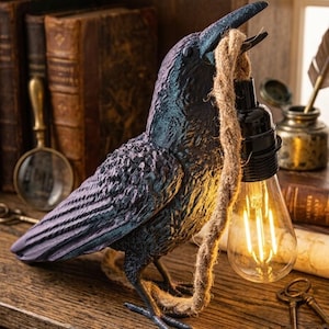 Può includere: Lampada decorativa a forma di corvo, con finitura strutturata e iridescente nei toni del blu e del viola. Il corvo tiene una lampadina nel becco, sospesa da una corda. La lampada è posizionata su una superficie di legno, con libri antichi e una lente d'ingrandimento sullo sfondo.