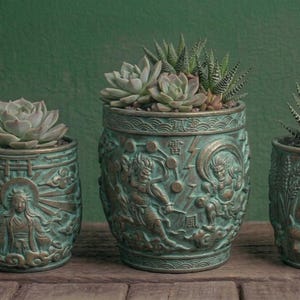 May include: Three cylindrical planters with a weathered bronze finish and relief carvings. The left planter features a deity, the center shows figures with lightning, and the right displays a fox mask. Each contains a green succulent plant.