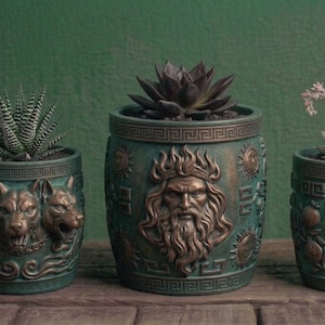 May include: Three cylindrical planters with a dark teal and bronze finish. The left planter features a three-headed dog, the center planter displays a bearded male figure, and the right planter shows a female profile surrounded by fruit motifs.