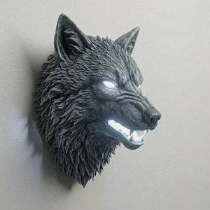 Könnte beinhalten: Graue Wolfskopfskulptur mit leuchtend weißen Augen und Zähnen, an der Wand befestigt. Die Skulptur hat eine detaillierte Fellstruktur und einen wilden Ausdruck, wodurch ein dramatisches Dekorationsobjekt entsteht.
