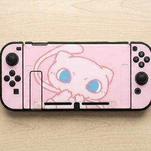 May include: A handheld video game console featuring a light pink skin with a stylized illustration of a pink creature with large blue eyes. The device includes black control sticks, directional buttons, and action buttons on the left and right sides.