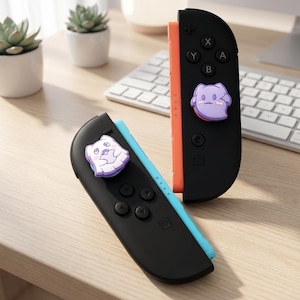 Op de afbeelding: Zwarte en oranje Nintendo Switch Joy-Con controllers met een paars en wit cartoon spook ontwerp. De controllers liggen op een houten oppervlak met een toetsenbord en kleine planten op de achtergrond.