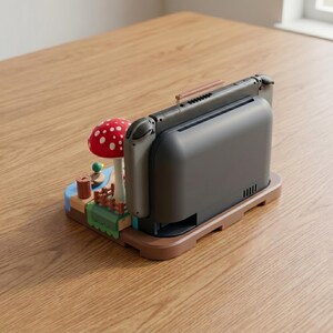 May include: A brown docking station for a gaming console, featuring a whimsical mushroom and landscape design. The console is dark gray, and the mushroom is red with white spots. The base is brown, and the landscape includes a small pond and fence.