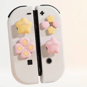 Puede incluir: Mandos Joy-Con blancos de Nintendo Switch con agarres decorativos para el pulgar en forma de estrella y flor en amarillo y rosa. Los mandos tienen detalles en negro y un botón más y menos.