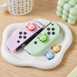 Peut inclure: Deux manettes de jeu aux couleurs pastel, une rose et une verte, avec des décorations d'animaux de dessin animé. Les manettes sont sur un plat blanc de forme ovale, avec des accessoires supplémentaires en forme d'animaux. Une tasse et une bougie décorative sont en arrière-plan.