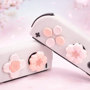May include: Two white game controllers with pink and peach-coloured accents. The controllers feature flower-shaped and circular buttons. The background is a soft pink with falling petals, creating a delicate design.