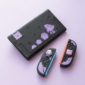 Puede incluir: Una funda negra para consola de juegos decorada con patrones de estrellas moradas y pegatinas de personajes, acompañada de dos mandos negros con detalles morados y agarres para pulgares en relieve con forma de personaje.