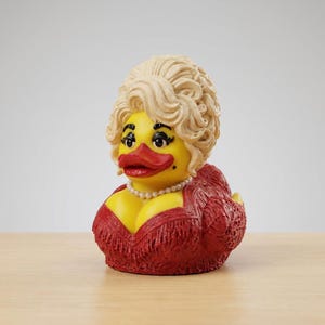 Puede incluir: Una figura de pato de goma amarillo con una voluminosa peluca rubia, un vestido rojo con flecos y textura de encaje, un collar de perlas blancas, pestañas oscuras y labios rojos. El pato tiene un pequeño lunar negro en la mejilla izquierda.