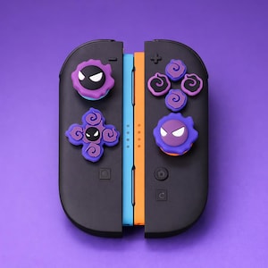 Może przedstawiać: Czarne kontrolery Joy-Con do Nintendo Switch z nakładkami na kciuki z motywem postaci w kolorze fioletowym, czarnym i białym. Kontrolery są umieszczone na fioletowym tle, z niebieskimi i pomarańczowymi szynami.