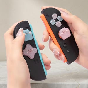 May include: Two black and colored Nintendo Switch Joy-Cons with Ditto-themed button covers. One Joy-Con is blue and the other is orange. The button covers are light blue and pink, with a glittery finish and smiling faces.