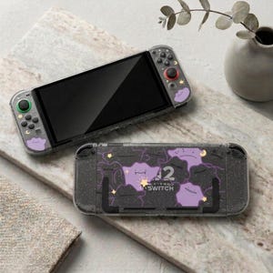 Könnte beinhalten: Eine transparente Nintendo Switch-Hülle mit einem schwarzen, gesprenkelten Design. Die Hülle zeigt lila Cartoon-Charakter-Grafiken und die Zahl 2. Die Switch liegt auf einer Marmoroberfläche.