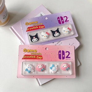 Op de afbeelding: Twee verpakkingen game console joystick caps voor een Switch. Eén verpakking bevat zwart-witte stripfiguren en de andere witte en roze figuren. De verpakking is roze en paars met de tekst "Game console joystick Cap".