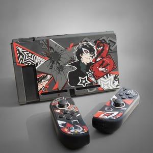 May include: A gaming console dock and two detachable controllers featuring a black, gray, and red graphic design. The artwork displays a character with a white mask, star patterns, and shattered glass effects, alongside red playing card motifs.