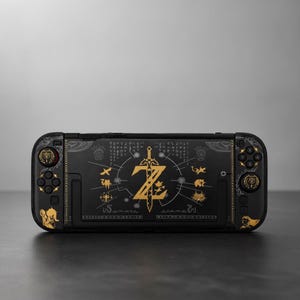 May include: A black handheld gaming console featuring gold-colored graphic designs. The center displays a large sword emblem with the letter Z, surrounded by various small icons, symbols, and intricate patterns across the device and controllers.