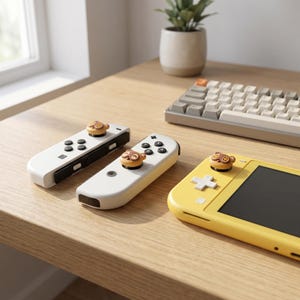 Op de afbeelding: Een gele Nintendo Switch Lite-console en twee witte controllers met bruine en tan duimgrepen met apen-thema worden getoond op een houten oppervlak. Een toetsenbord en een kleine plant in een pot zijn ook zichtbaar.