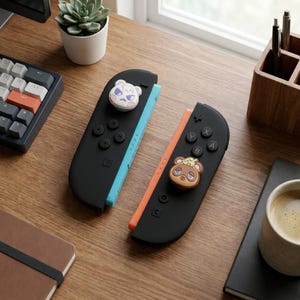 May include: Two black Nintendo Switch Joy-Cons with character-themed button covers. One Joy-Con has a blue side and a white bear button cover. The other has an orange side and a brown bear button cover. The controllers are on a wooden surface.