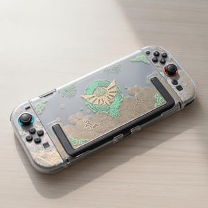 May include: A handheld gaming console featuring a clear protective case with gold and mint green intricate patterns. The design includes a central emblem, swirling circular motifs, and geometric shapes across the back and controller sides.